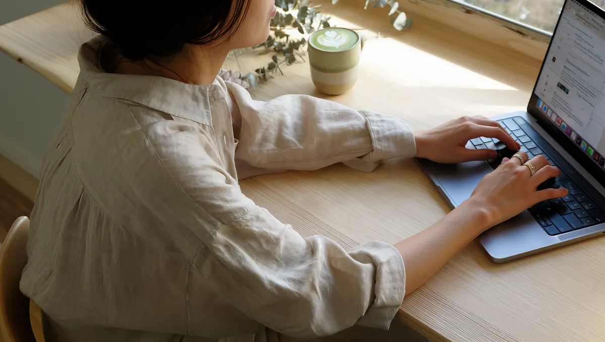 Over-the-shoulder view of a woman typing on a laptop at a sunlit oak desk